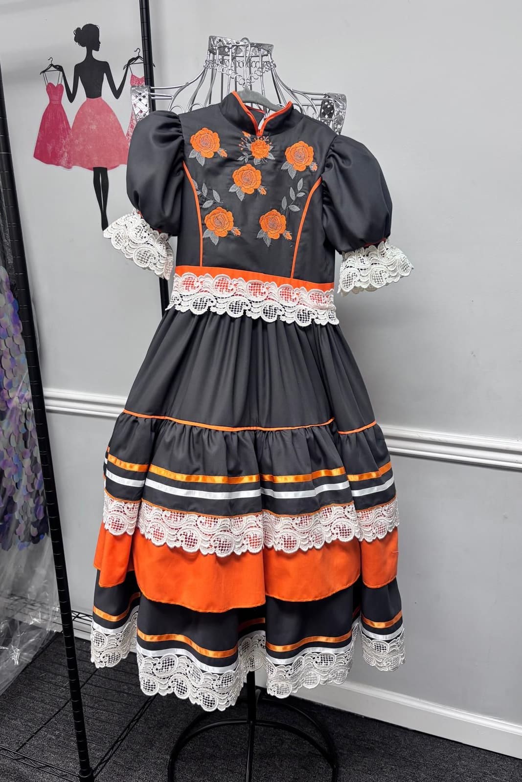 Black folkloric dress with orange embroidered roses, puff sleeves and tiers of lace and ribbon on a stand
