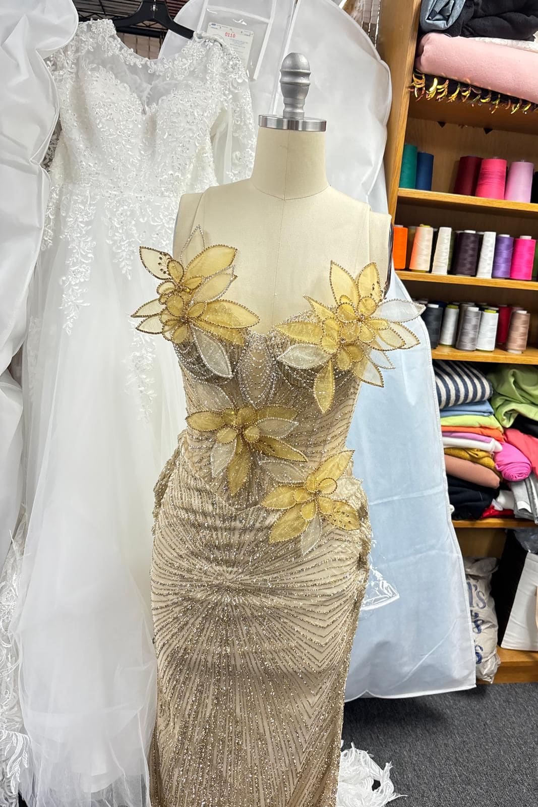 Champagne beaded gown with hand-shaped gold organza flowers across the bodice, on a dress form in the atelier